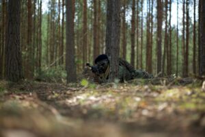 Soldier camouflaged in a forest environment, holding a weapon in a tactical stance.